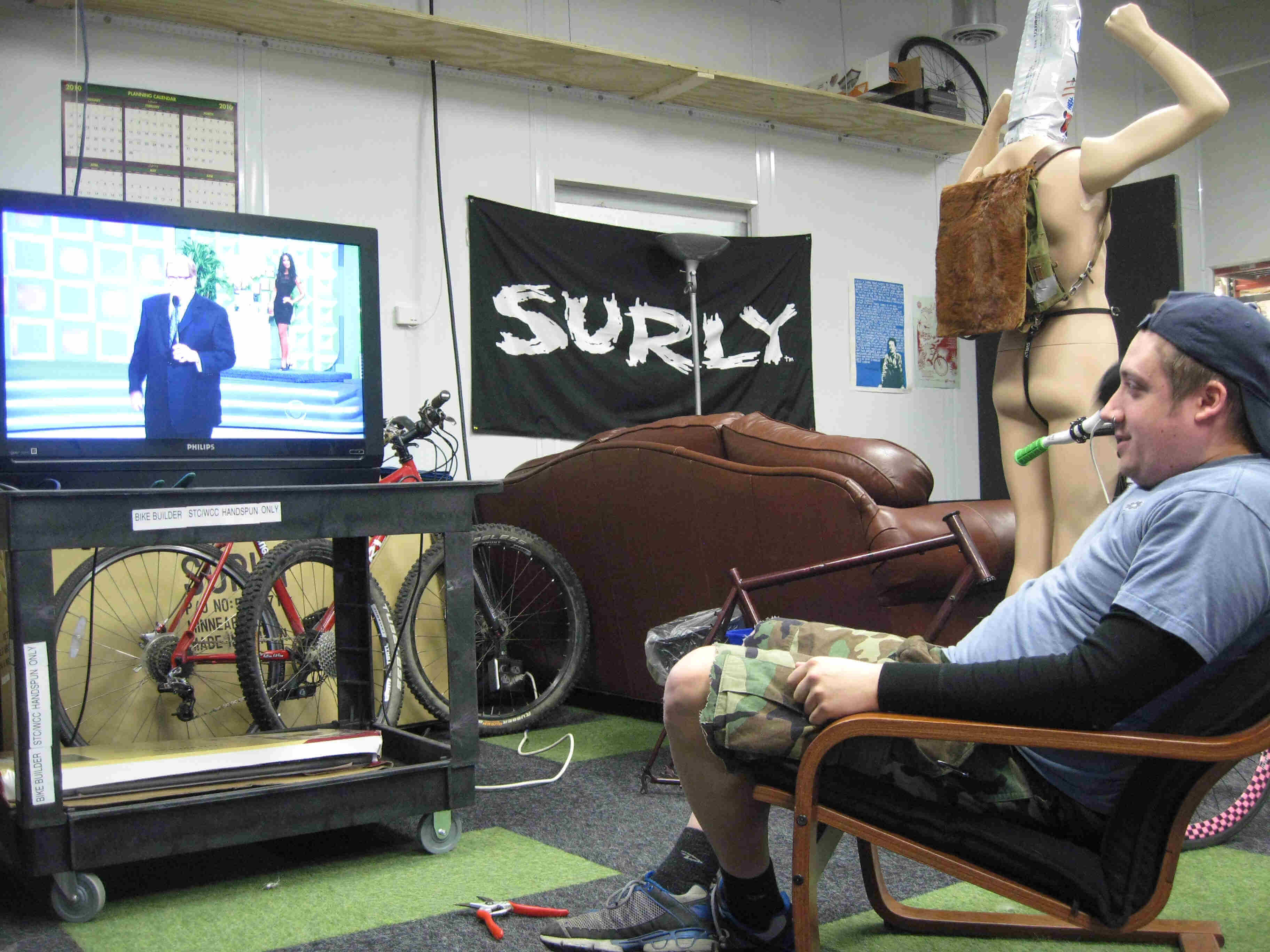 Left side view of a person sitting in a chair, watching a TV on a cart, in a room with a Surly banner on the wall